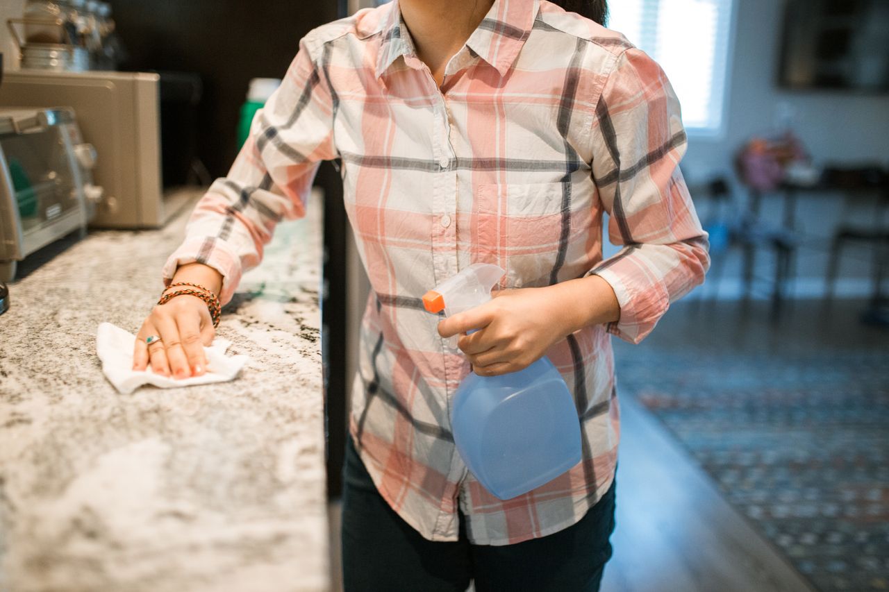 services-01 A woman wearing a plaid shirt cleans a kitchen counter using a spray bottle and cloth.